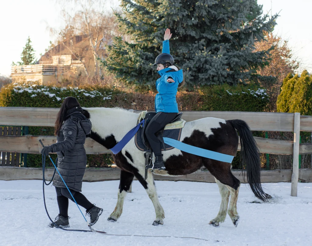 Longenführerin begleitet Reiterin auf einem schwarz-weiß gescheckten Pferd bei einer Übung im Schnee.